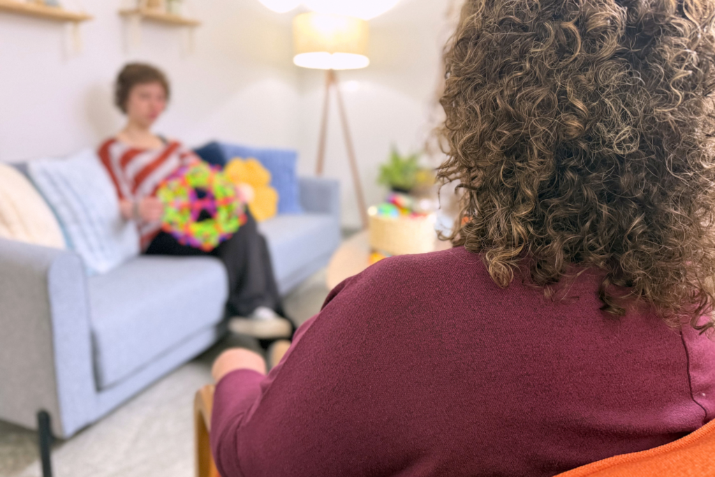 Over a therapists shoulder blurred in the background you can see a teenager holding a fidget toy and playing.