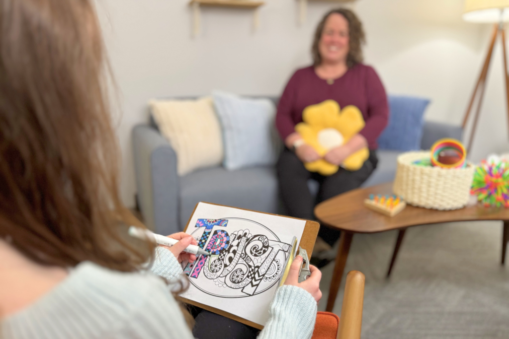 Over the shoulder of a kid colouring a sheet that reads "trust" a therapist can be seen smiling in the background.